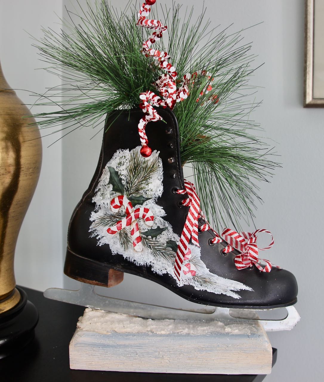 Closeup of one vintage ice skate om a block of wood with faux snow. Painted on the side of the skate is some fir branches and candy canes with fir branches flowing from the top of the skate