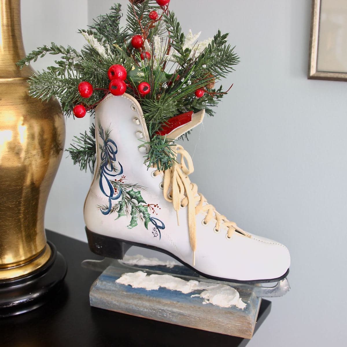 Closeup of a white vintage ice skate on a block of wood with faux snow at the bas of the blade, holly painted on the side of the boot and grrens with berries flowingout the ankle of the skate