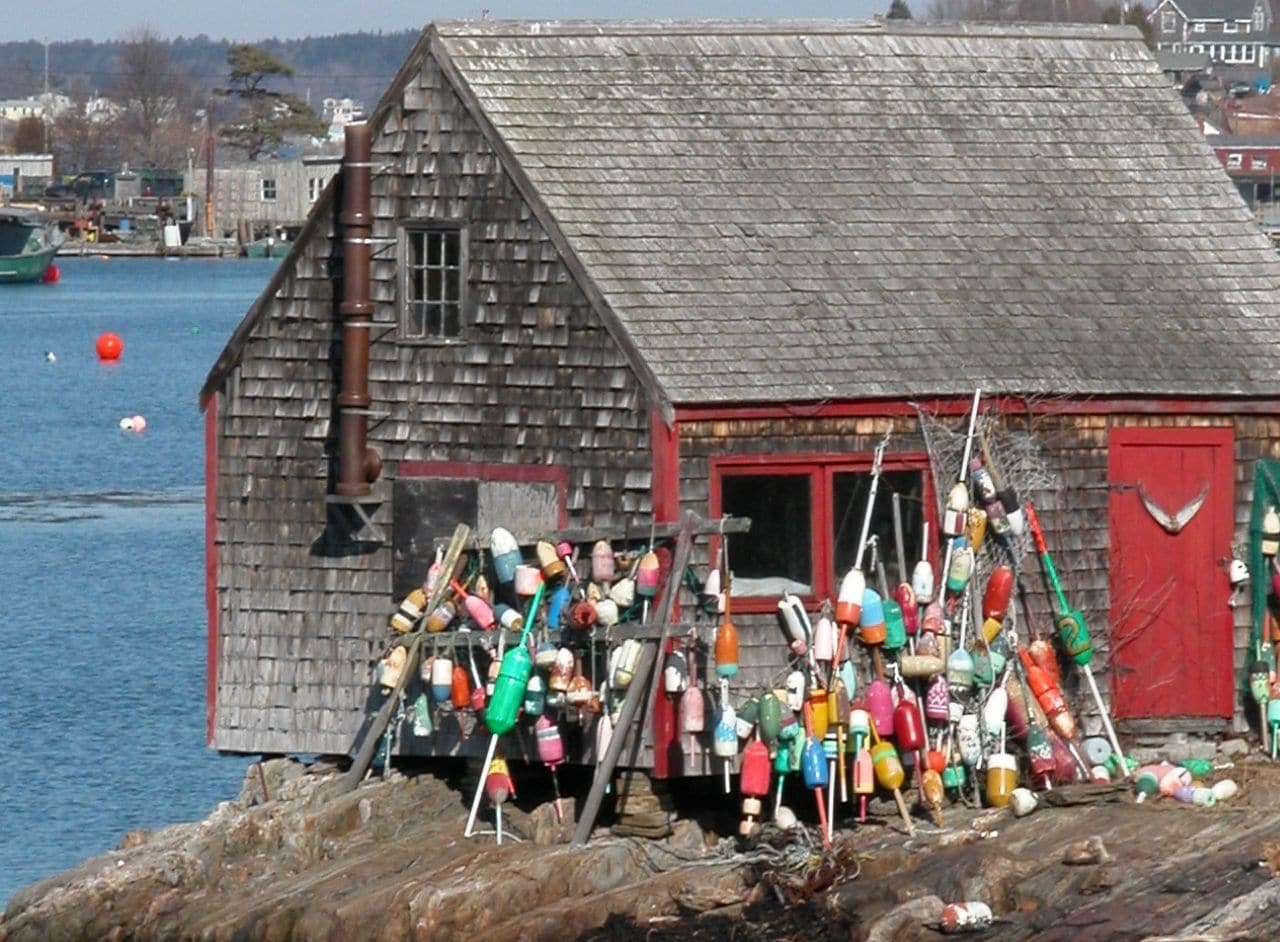 The side of a weathered building on the ocean with many many old wooden buoys piled up and hanging on one side of the house