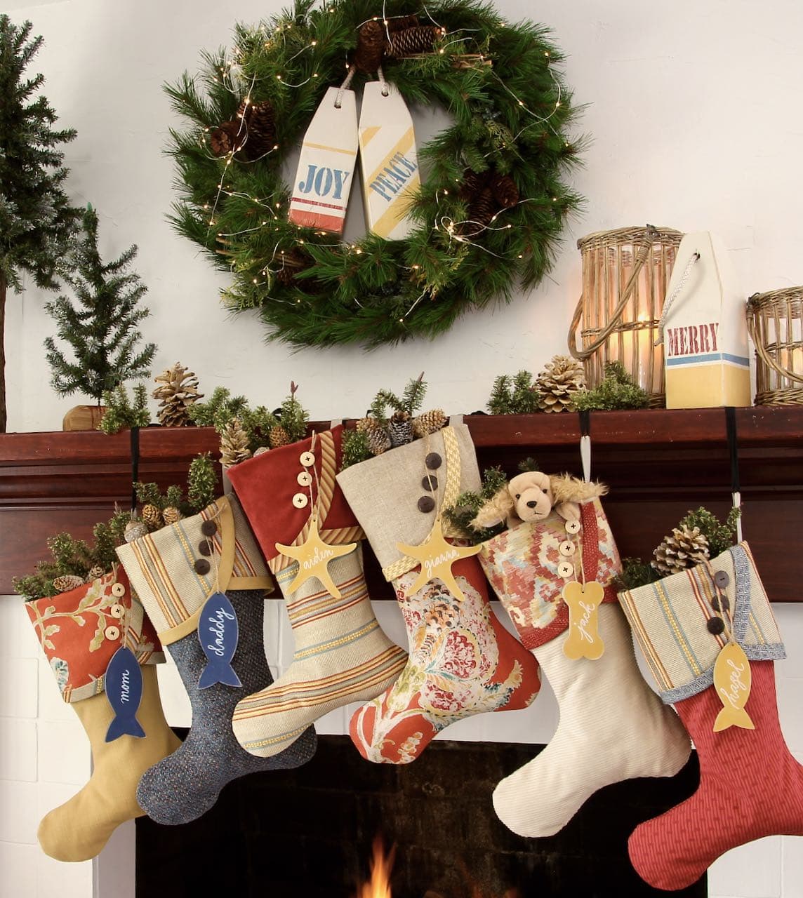 full view of the fireplace with six Coastal Christmas stockings hanging from a wood mantel with a wreath above with two Christams buoys in it and a third sitting on the mantel with rattan hurricane candleholders and skinny woodland trees and pinecones