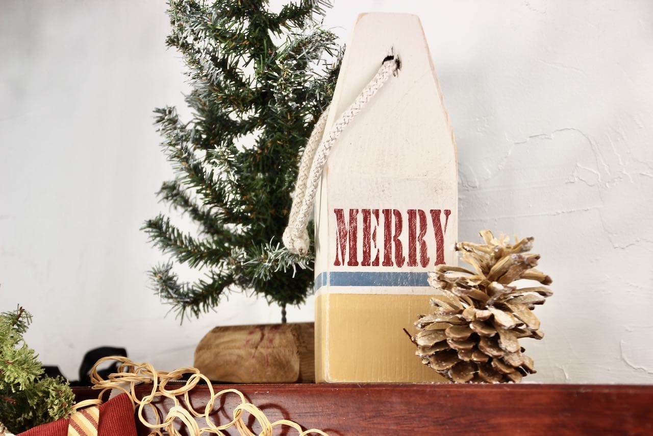 Closeup of "Merry" buoy on corner of wood mantel with a small fir tree behind one side and a bleached pinecone in front on the other side