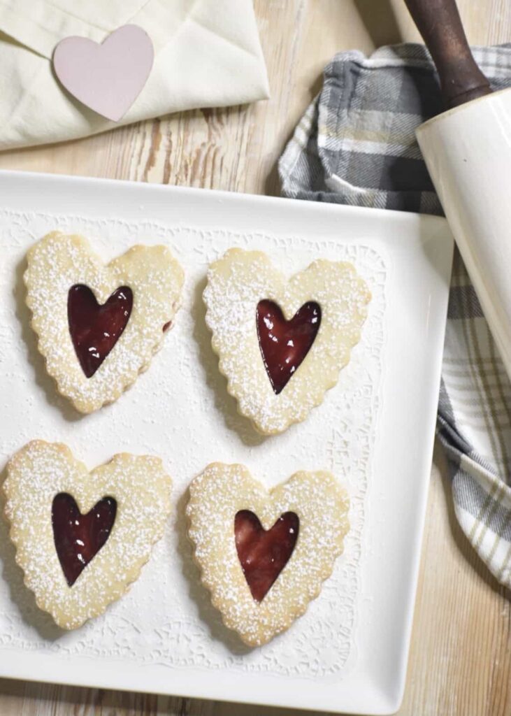 Part of a serving tray with four heart-shaped Linzer cookies  and a tea towel. and rolling pin off to the side