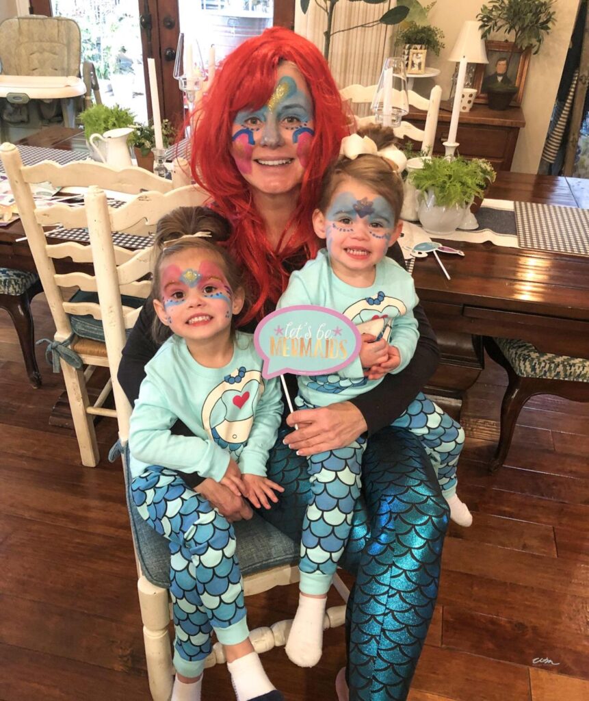 two young girls dressed up as mermaids on the lap of a woman also dressed up as a mermaid
