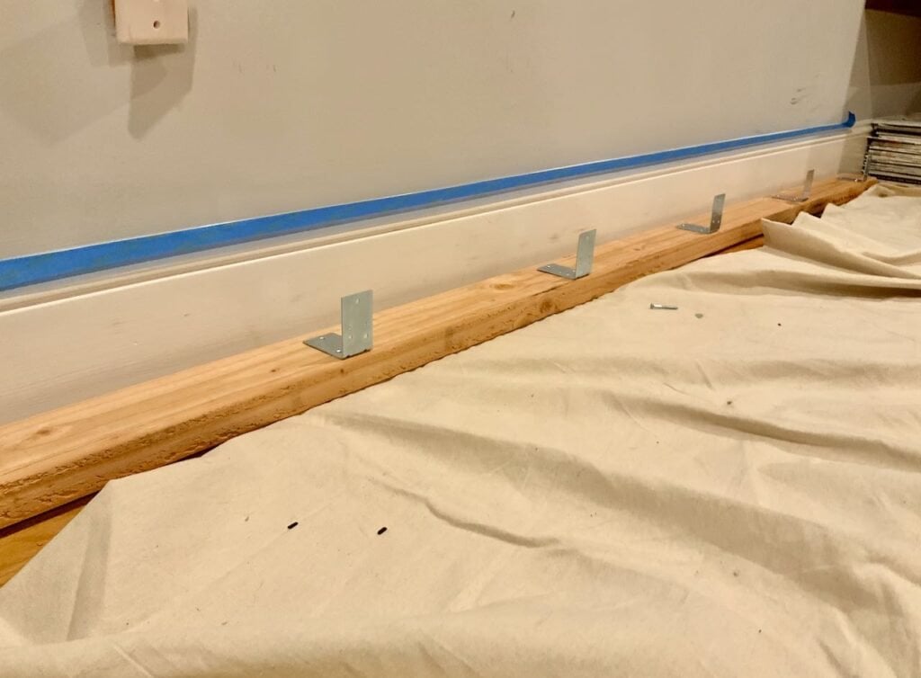 2 x 4 is shown laying next to the base moulding with L brackets shown lined up with the wall stud locations