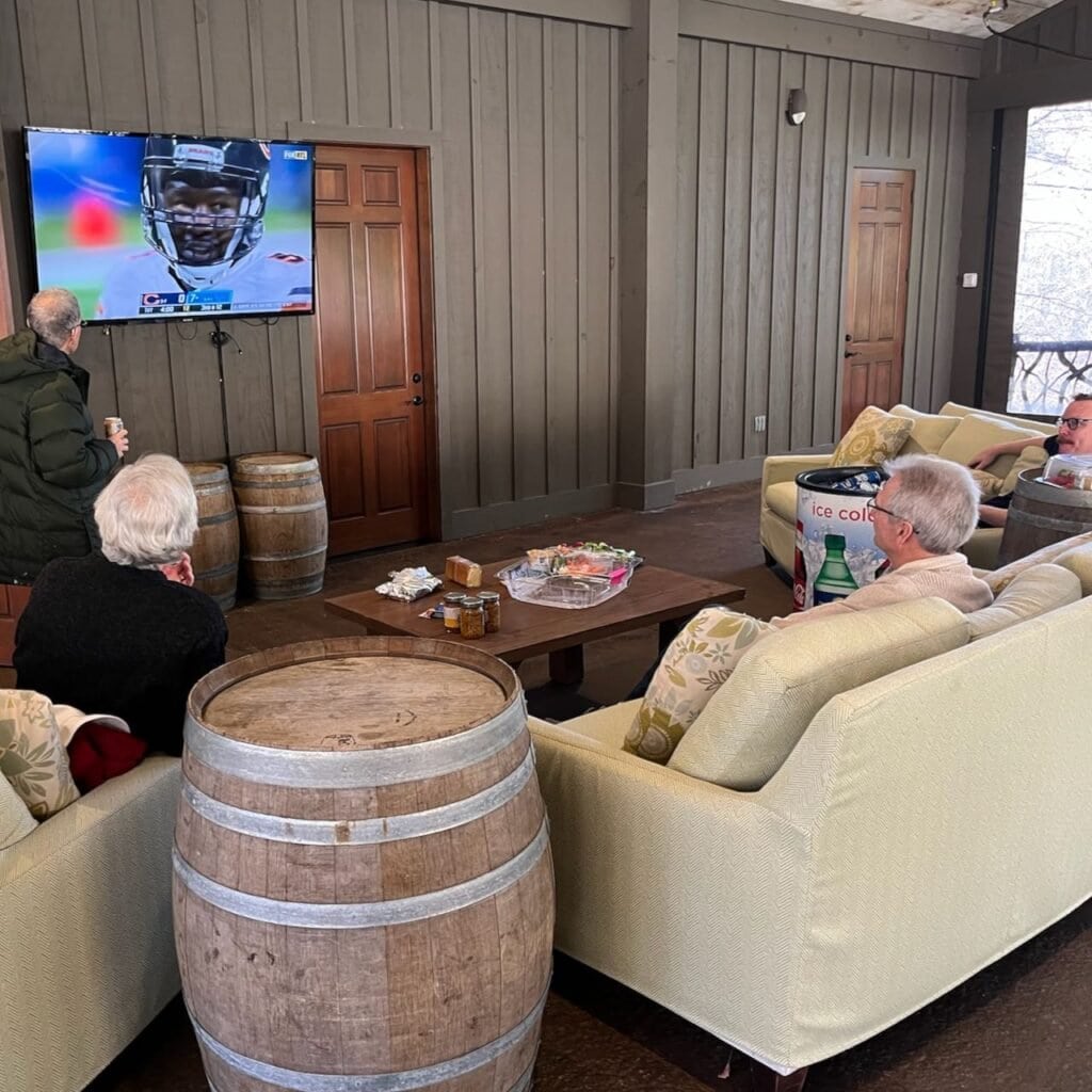 Several men sitting on and standing by a pair of couches watching a football game on a big screen TV on Thanksgiving
