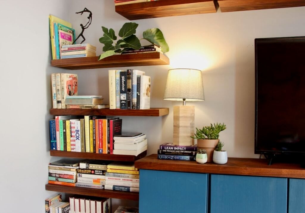 One corner of blue cabinet with walnut countertop with walnut floating bookshelves beside and an accent light and trio. of plants on the counter 