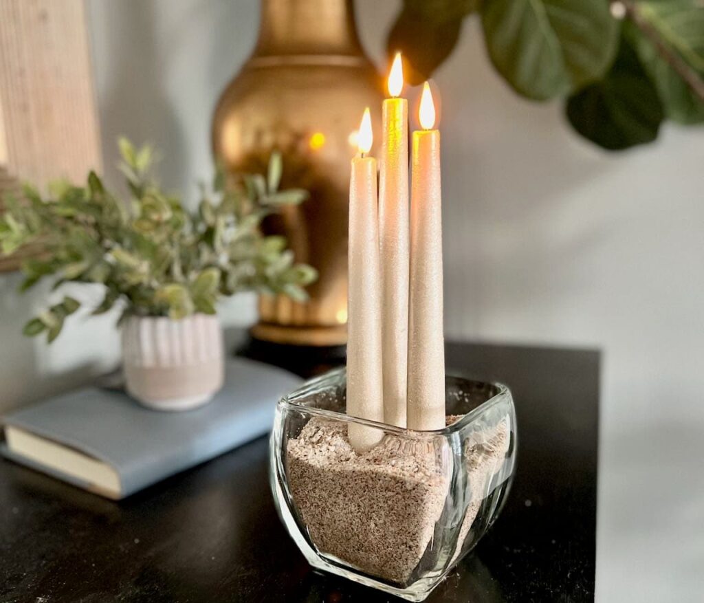 Three vintage gold battery operated taper candles are lit and standing in a glass bowl filled with sand