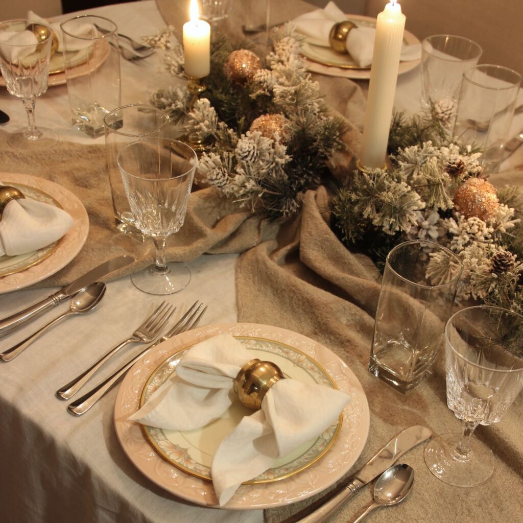 Candlelit scene of a white linen tablecloth with linen table runners criss crossing in the center of the centerpiece arrangement with rose gold decor and each place setting has a blush dinner plate with an ornate salad plate with a white napkin folded as a bow with a brass napkin ring