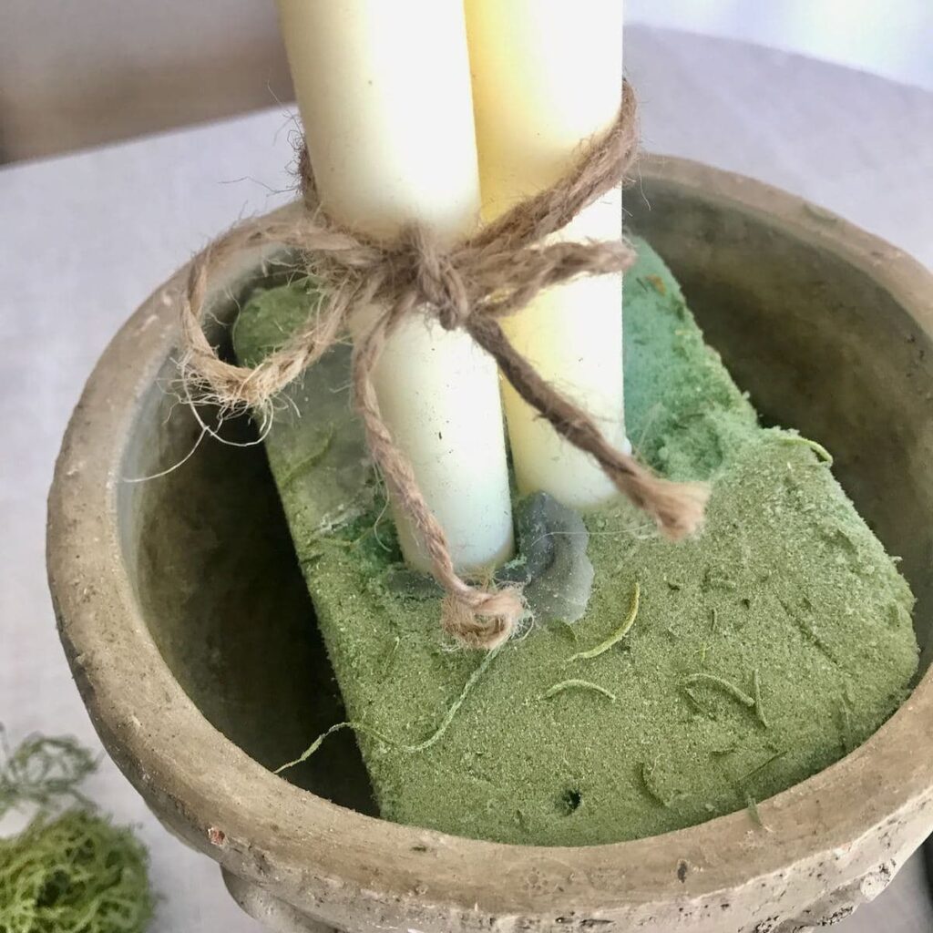 A bundle of three white taper candles are held together with twine tied in a small bow. Then they are held upright by florist foam tucked inside a concrete urn