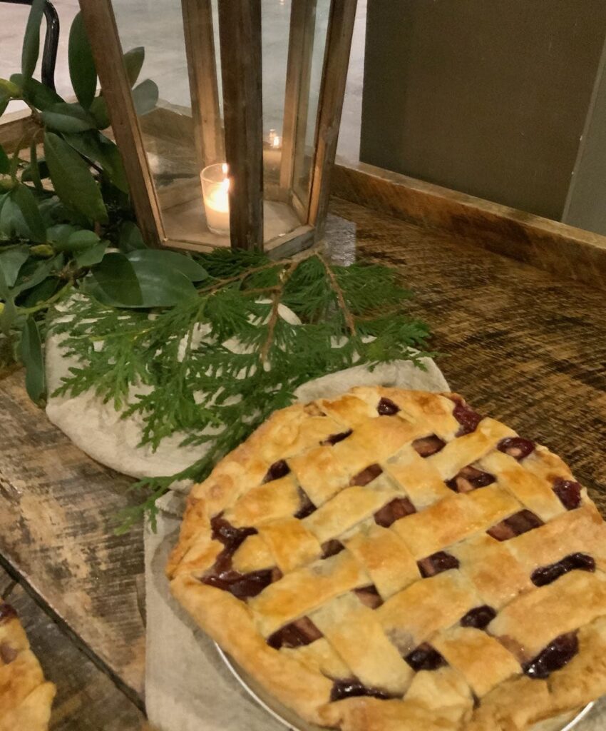 Another vintage table adorned with a. large vintage lantern with votive candle and greens and a washed linen table runner with a freshly baked pie