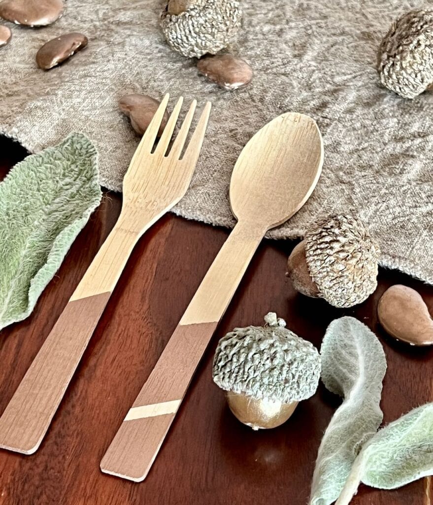 A bamboo fork and spoon with the handles painted a bronze metallic laying by a washed linen table runner, some dried lambs ear and some acrorns and lima beans also painted in metallic bronze and champagne