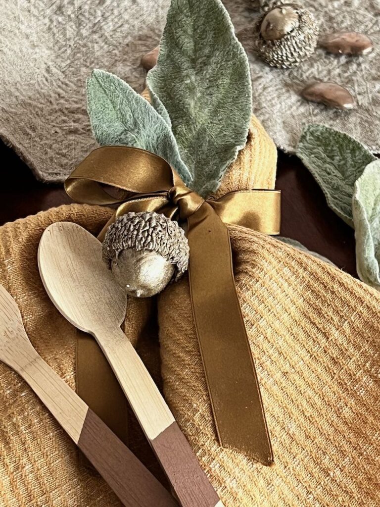 Closeup of a napkin tied with a satin ribbon with a bamboo spoon and fork, washed linen table runner, dired lambs ear and acorns