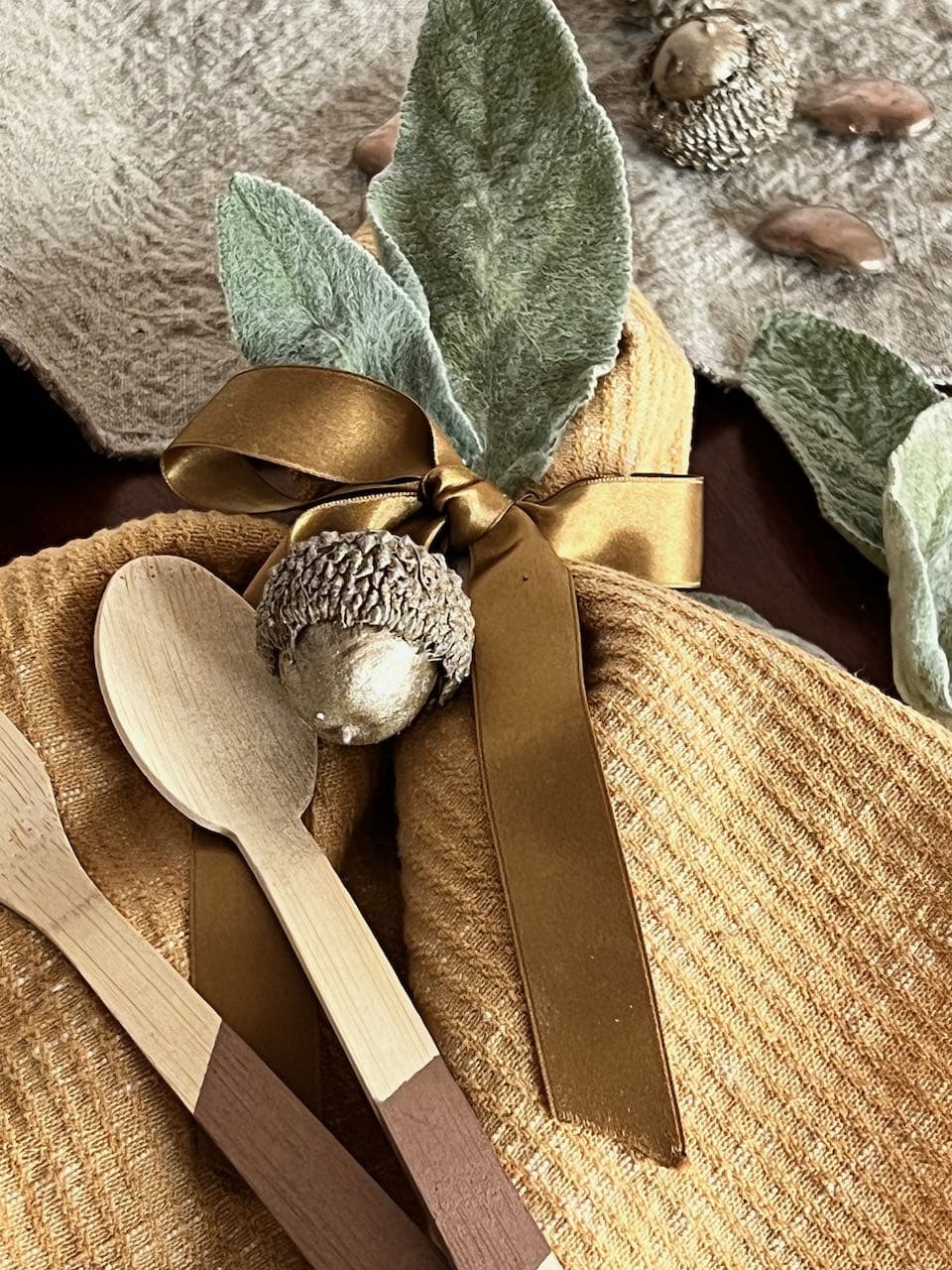 Closeup of a napkin tied with a satin ribbon with a bamboo spoon and fork, washed linen table runner, dired lambs ear and acorns