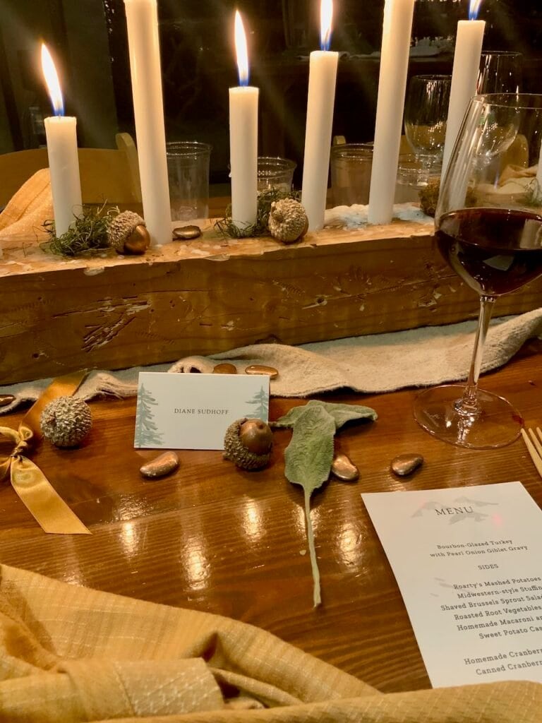 Friendsgiving Table decor of a distressed wood block is holding 6 lit candles, some dried moss, bronzed acorns and lima beans, with a napkin, ribbon and lambs ear, a place card and menu