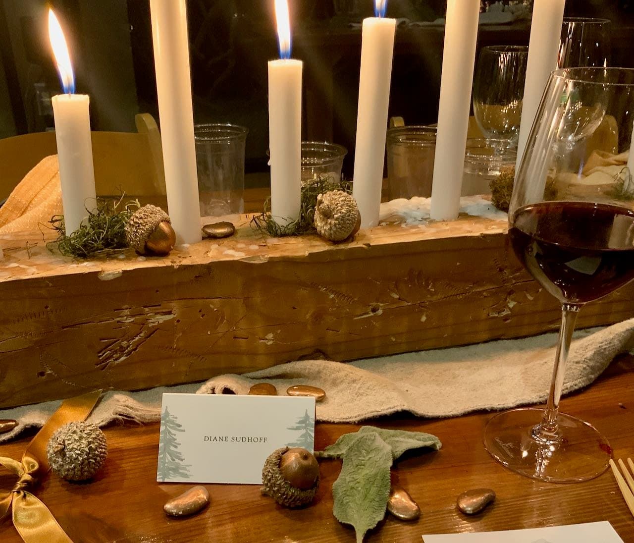 Lit candles in a distressed wood block with scattered bronzed acorns, lima beans, a napkin, partial wine glass and a place card
