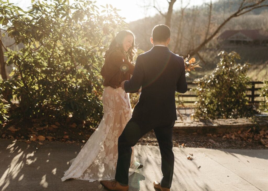 A groom with his back to us is hold the brides flowers and her hand helping her down a wooded walkway