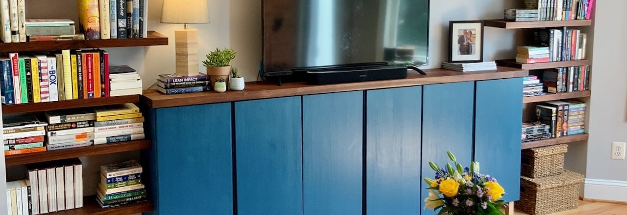 Blue wall mounted cabinets with a walnut countertop holding a Tv and some plants and a framed photograph, surrounded with floating walnut bookshelves