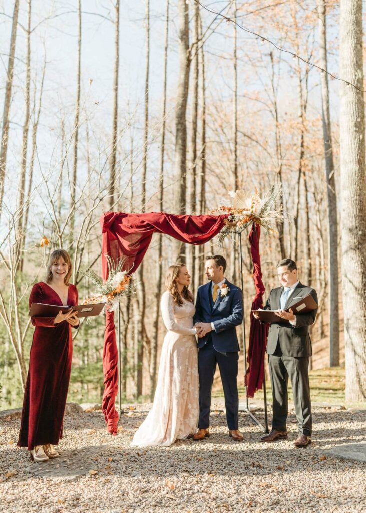 under a blue sky, long view of a bride and groom with two officiants standing in a chuppah wrapped in gauze with two dried floral arrangements