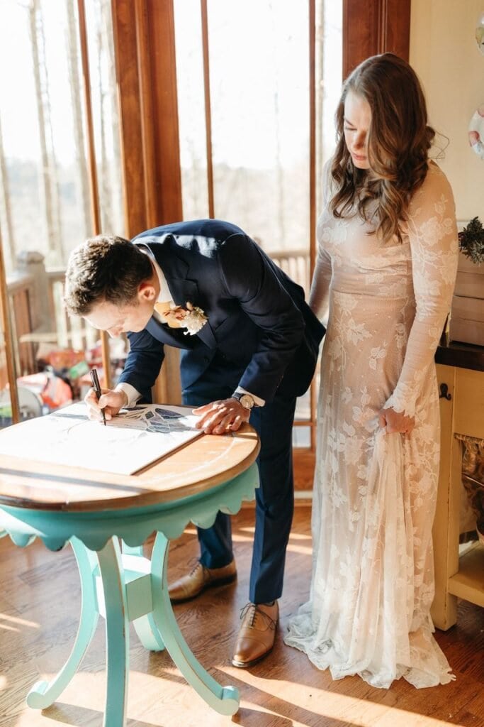 Groom is bent over small round table with bride next to him while he signs the ketubah