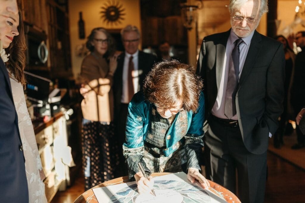 Groom's mother is shown signing the Ketubah whil his father stands by and the brides parents are in the background