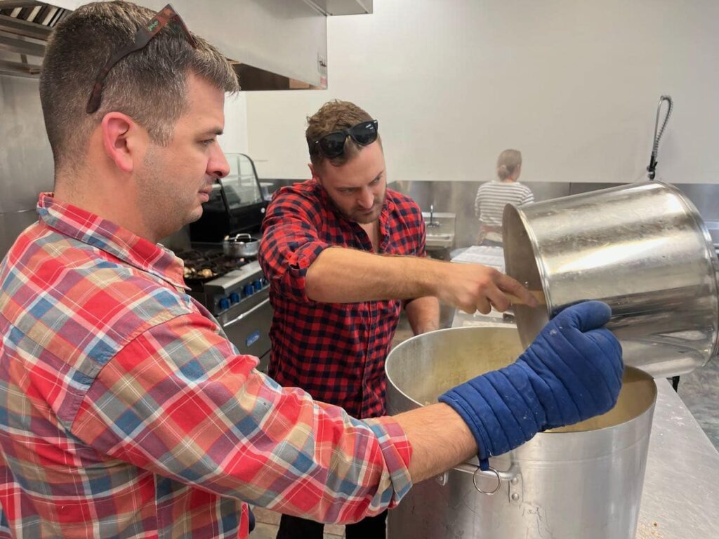 Two young men in plaid shirts are pouring a large pot's contents into an even larger pot