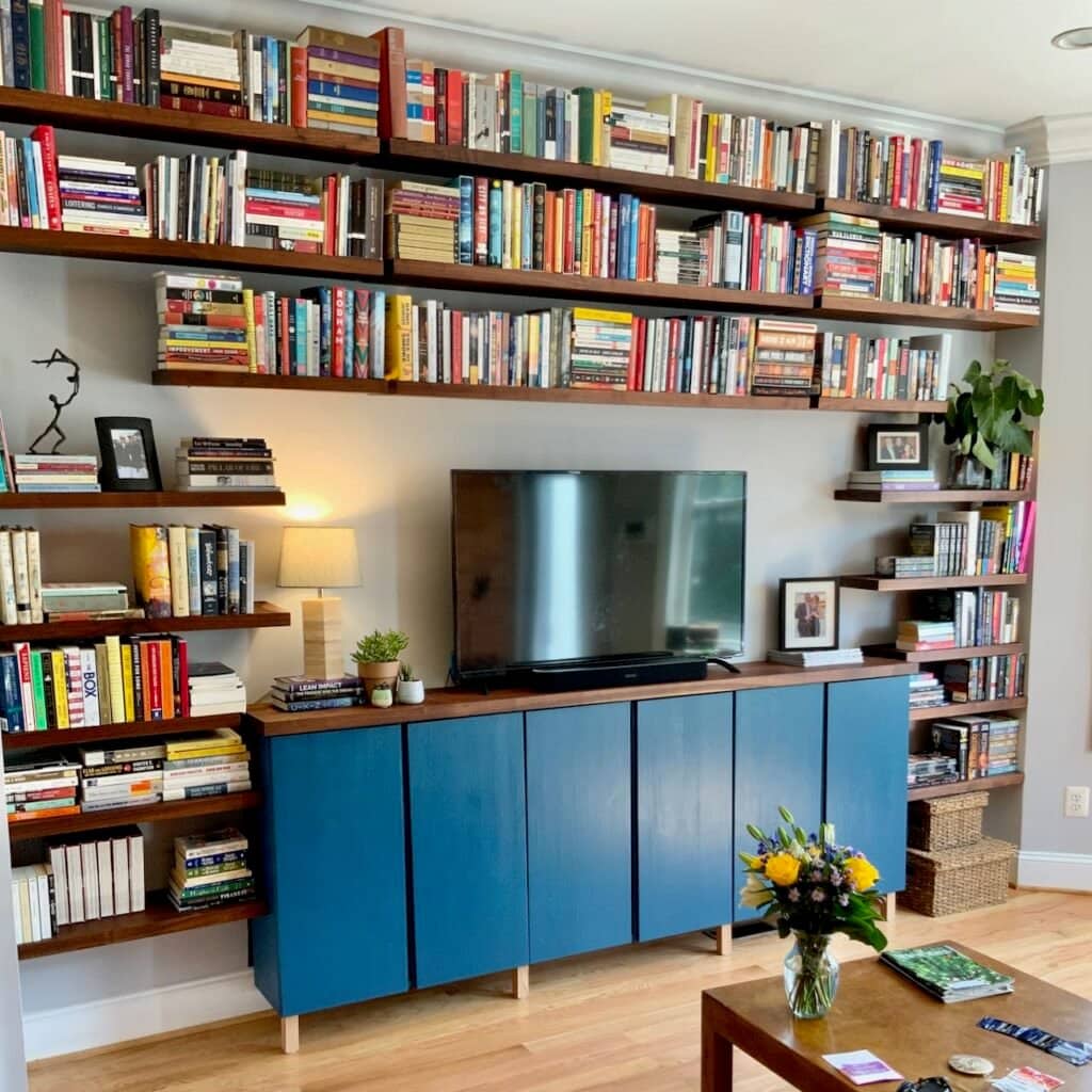 Beautiful living room wall filled floor to ceiling with floating book shelves surrounding three deep blue cabinets with a TV on top, accent lamp and plants