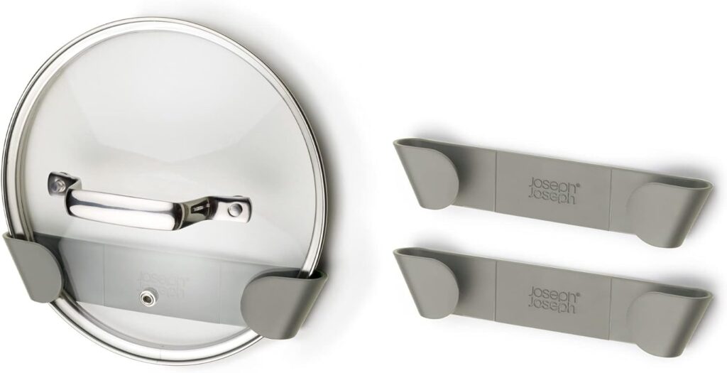 A glass pot lid is held vertically by two gray plastic wall-mounted holders labeled "Joseph Joseph," making it a neat solution for organizing cookware in a walk-in pantry. A second empty pair of holders is shown beside it.