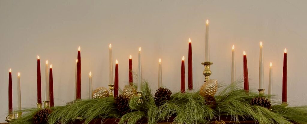 A wood mantel is covered in green pine branches with pinecones attached. Down the length of the mantel are 23 mismatched brass candlesticks holding a mix of burgundy and vintage gold battery operated candles