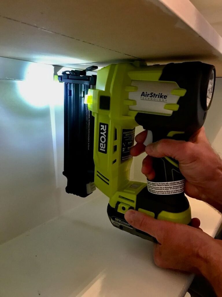 A man's hands are holding a Ryobi nail gun right against a furring strip attaching it to the inside of the pantry back wall.