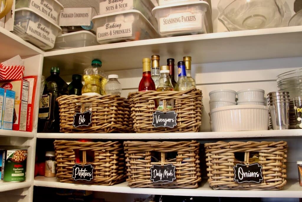 Pantry shelves loaded with clear plastic boxes labeled "Baking: Nuts & Such", Baking: wrappers, Straws, Etc", "Seasoning Packets' etc. And on two lower shelves are woven baskets with chalkboard labels of "oils", Vinegars". "Potatoes", "Onions" and "Daily Meds"