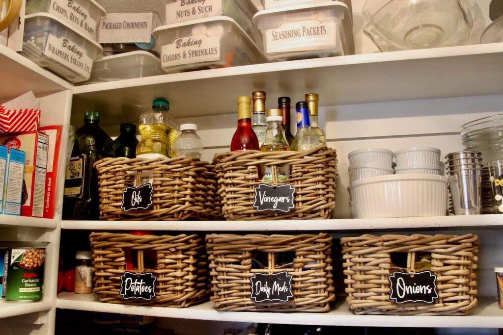 Pantry shelves loaded with clear plastic boxes labeled "Baking: Nuts & Such", Baking: wrappers, Straws, Etc", "Seasoning Packets' etc. And on two lower shelves are woven baskets with chalkboard labels of "oils", Vinegars". "Potatoes", "Onions" and "Daily Meds"