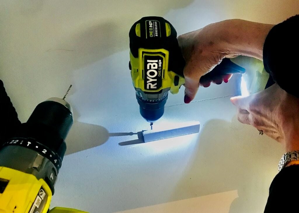Woman's hand operating a Ryobi hand drill with a Phillips head bit, driving a screw into the Platter lip, attaching it to the lowest pantry shelf