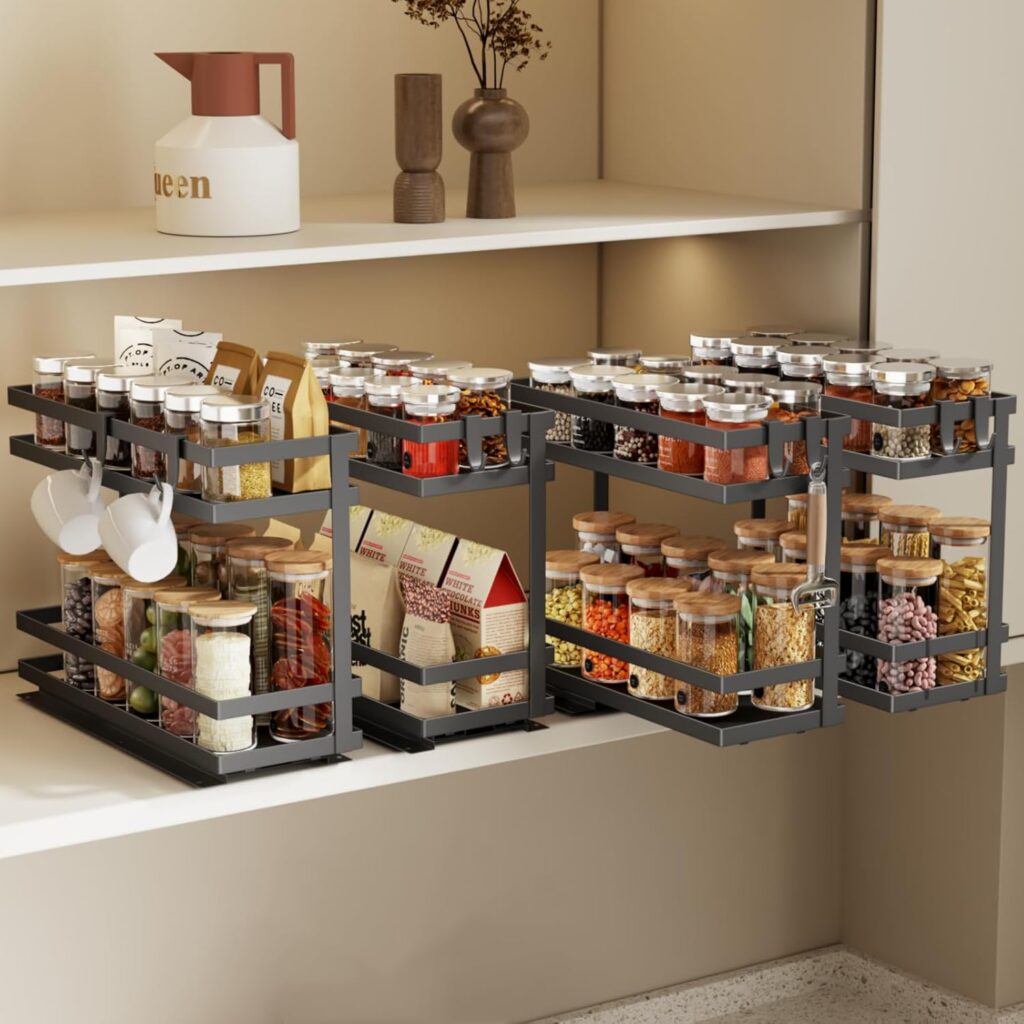 A well-organized kitchen shelf with black pull-out racks holding spice jars with labels, cans, packets, and spices, all neatly arranged in transparent containers. White mugs hang from the left rack, and decorative items sit on the upper shelf.