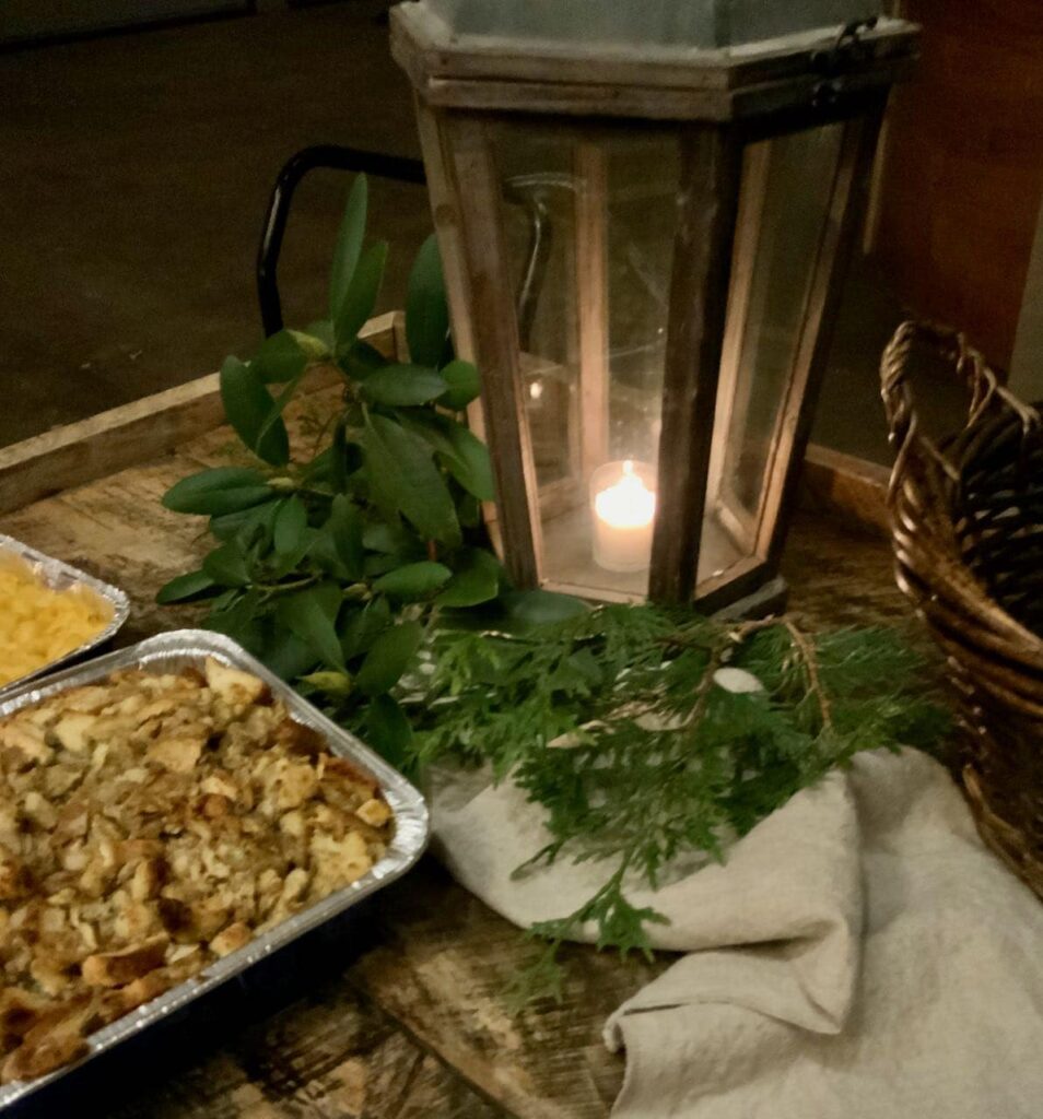 Vintage table decorated with a large vintage lantern holding a votive candle with magnolia greens and a washed linen table runner that holds a third serving dish of all the side dishes