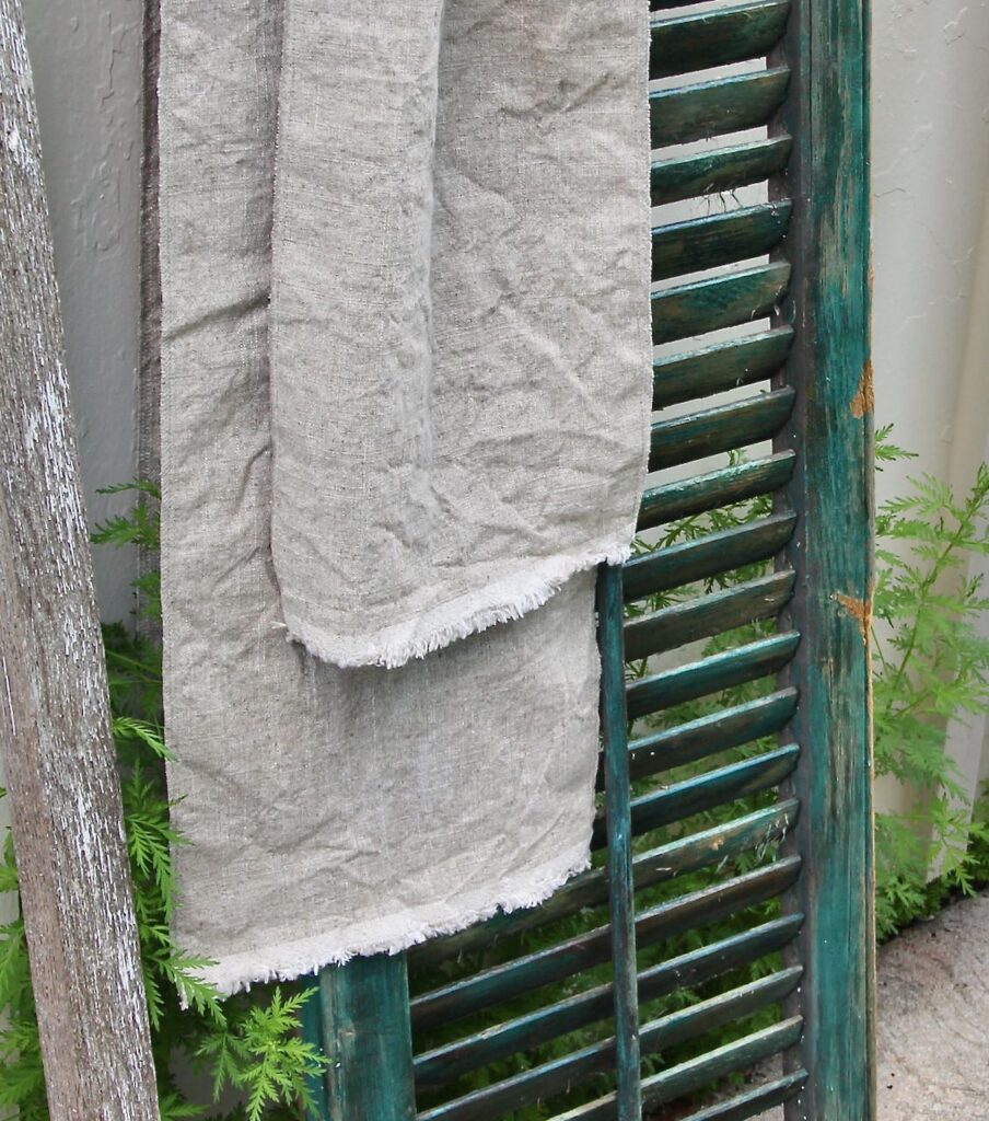 Two Skinny Washed Linen Runners with fringed ends draped over a vintage shutter with plants growing up behind