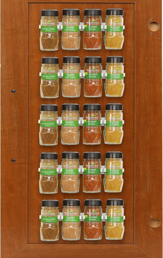 A wooden spice rack mounted on a door, holding 18 spice jars with labels in green and white, arranged neatly in three columns and six rows.