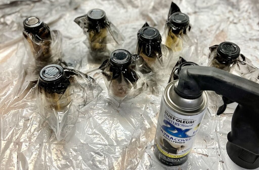 Spice bottles in sandwich bags with the lids shown painted black sitting next to a can of black spray paint with a pistol grip spraying attachment