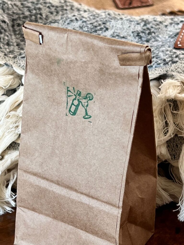 Brown craftpaper treat bag with the top folded down and a stamped with their wedding logo of a beer bottle clinking a margarita glass