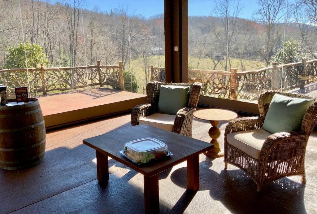 Two comfy chairs and table in front of floor to ceiling clear wall overlooking the view of the Blue Ridge. Mountains