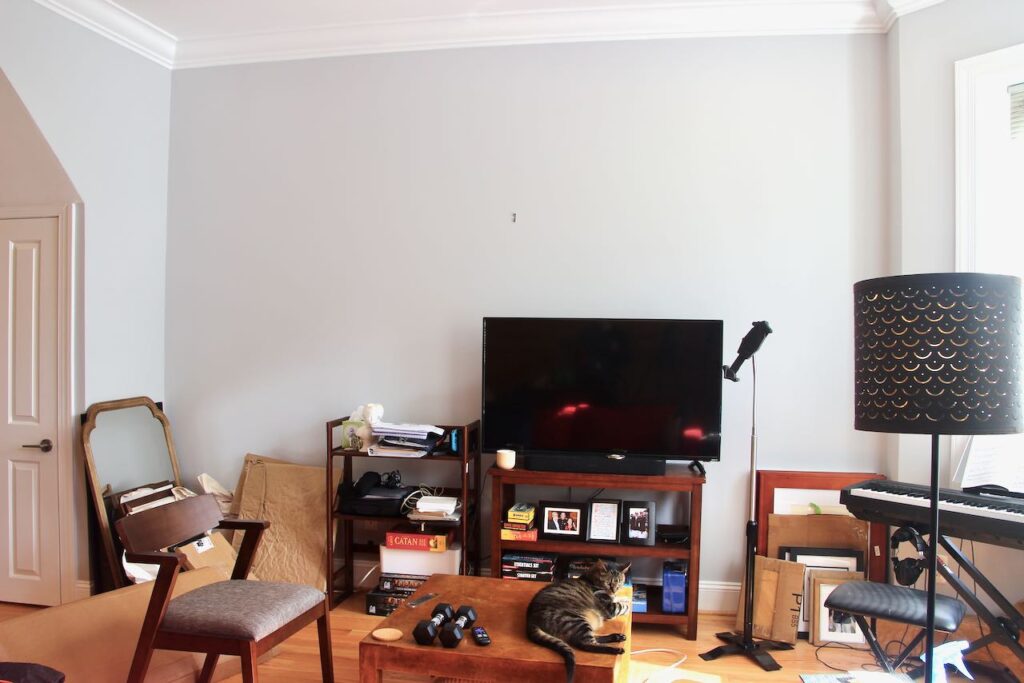 A tall light colored wall with piles of artwork, lamps, a tv, keyboard and misc moving debris