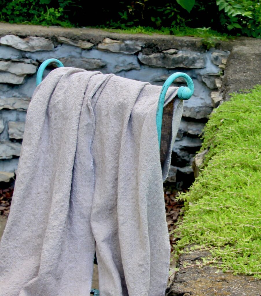 Two wide Washed Linen Table Runners draped over a vintage Garden Chair in front of a stacked stone wall with ferns and ground covering