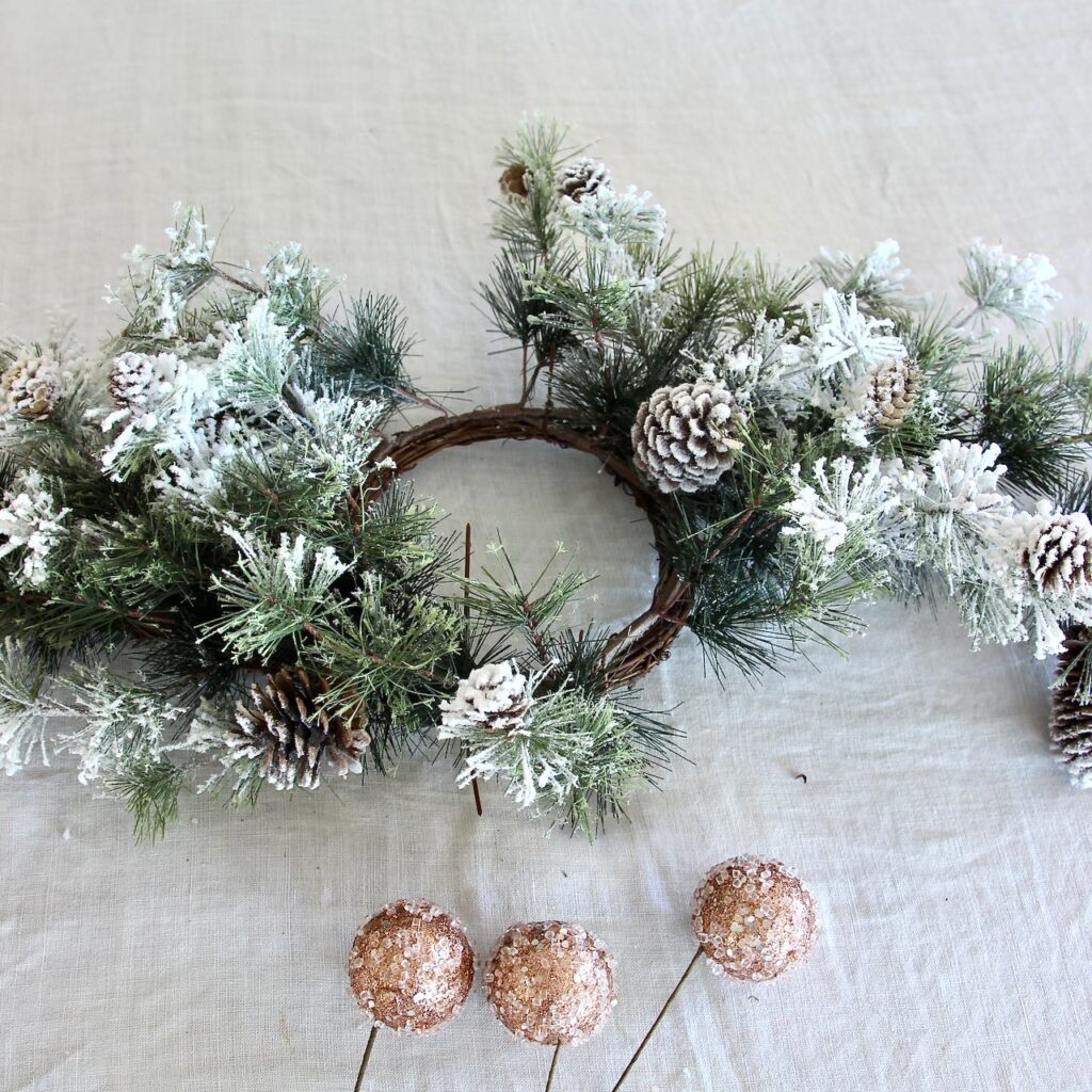 A candle ring of willow branches with flocked fir branches and pinecones and three rose gold bulbs on wire picks