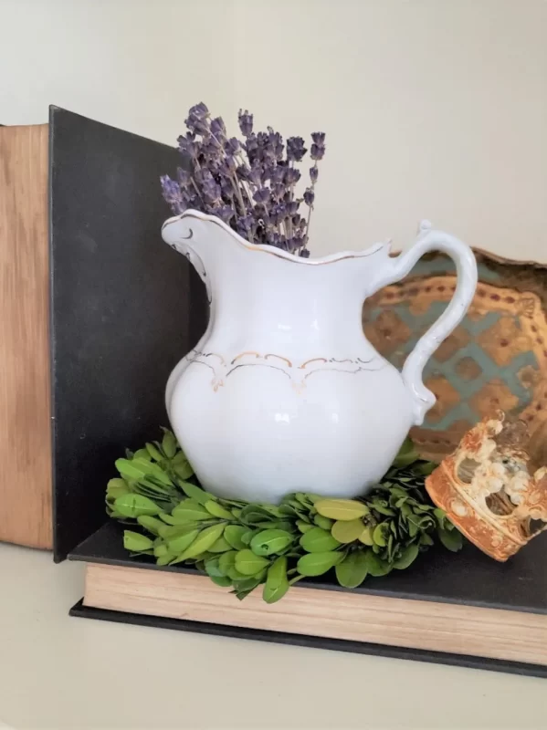 A vintage white pitcher filled with dried lavender sitting on a boxwood wreath on a book with aged pages