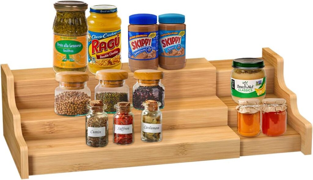 A tiered wooden spice rack holds spice jars with labels, along with two jars of peanut butter, a jar of pasta sauce, a jar of pickles, a jar of applesauce, and two small jars of orange preserves.