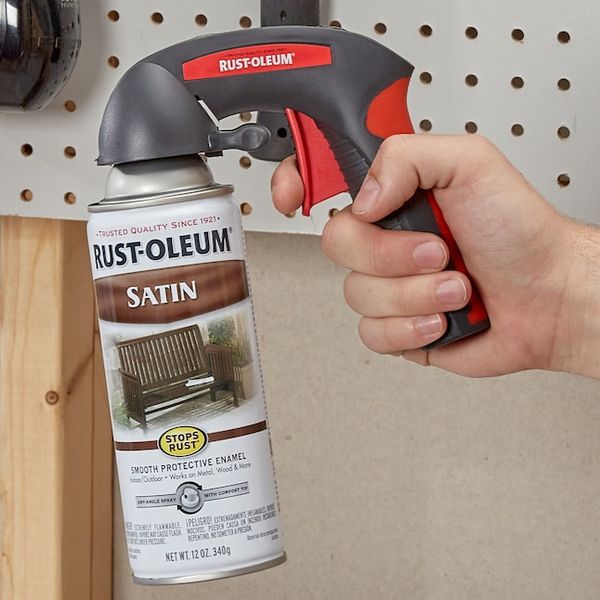 A hand holds a Rust-Oleum satin spray paint can with a red and black comfort grip trigger, aimed at a pegboard wall in a walk in pantry or workshop setting.