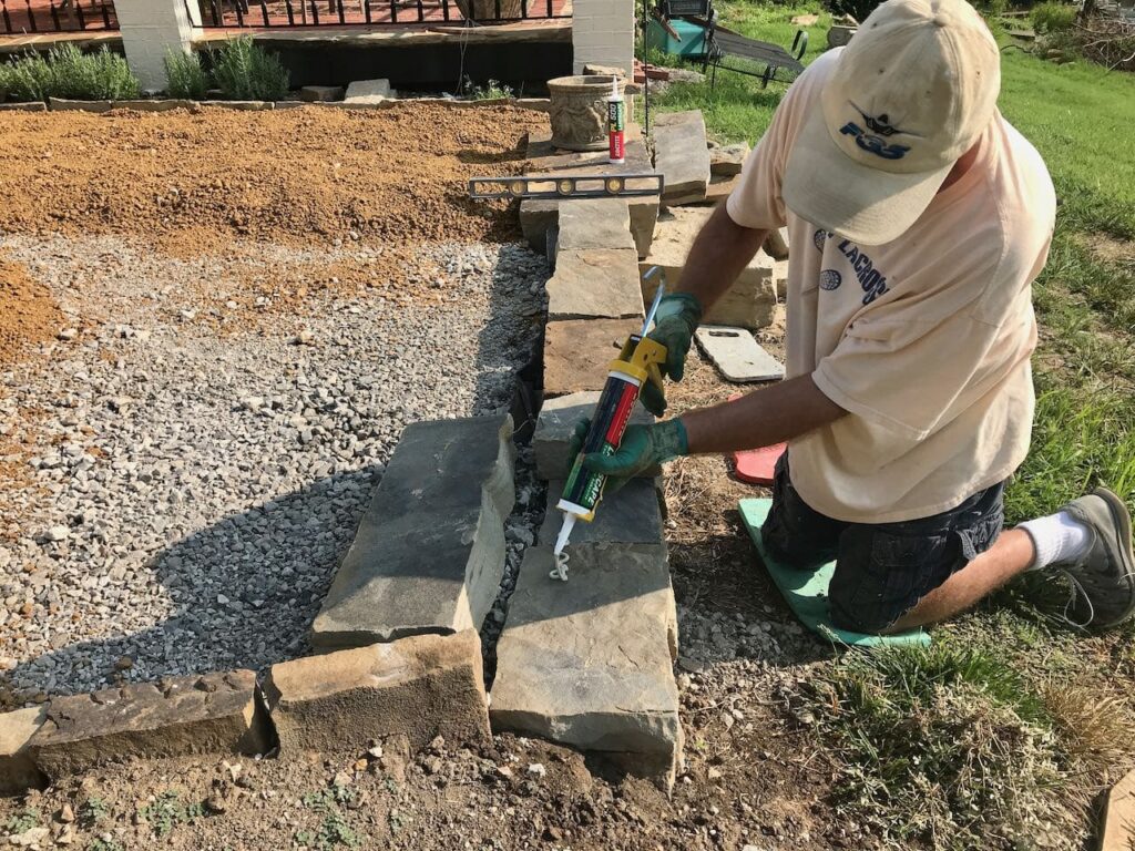 Man kneeling by the retaining wall applying adhesive to the last three top layer stones. You can see the road fill gravel in place and some of the DG in the background 