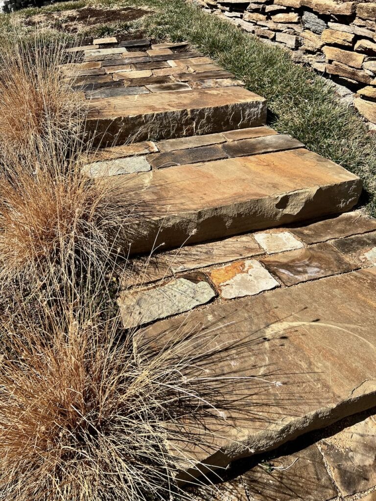 View of three finished stone steps with flagstone extended landings between them and tufts of dried ornamental grass on the side marking each step