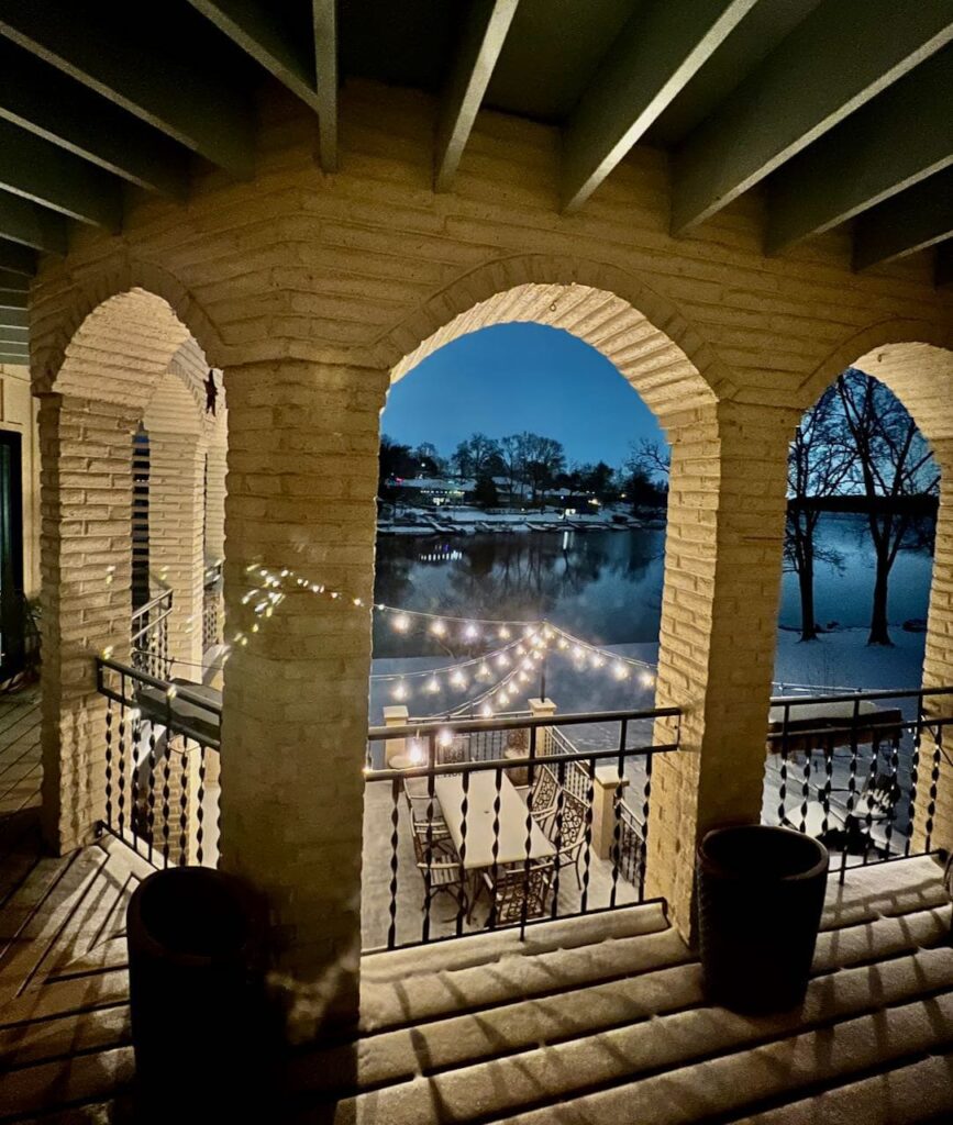 view from the large balcony above, through a large brick archwayof a snow-covered patio and balcony floor and railings all lit by the four strands of outdoor cafe lights suspended from the brick columns of the balcony out to a metal pole on. the corner of the patio below. All with a lake in the distance.