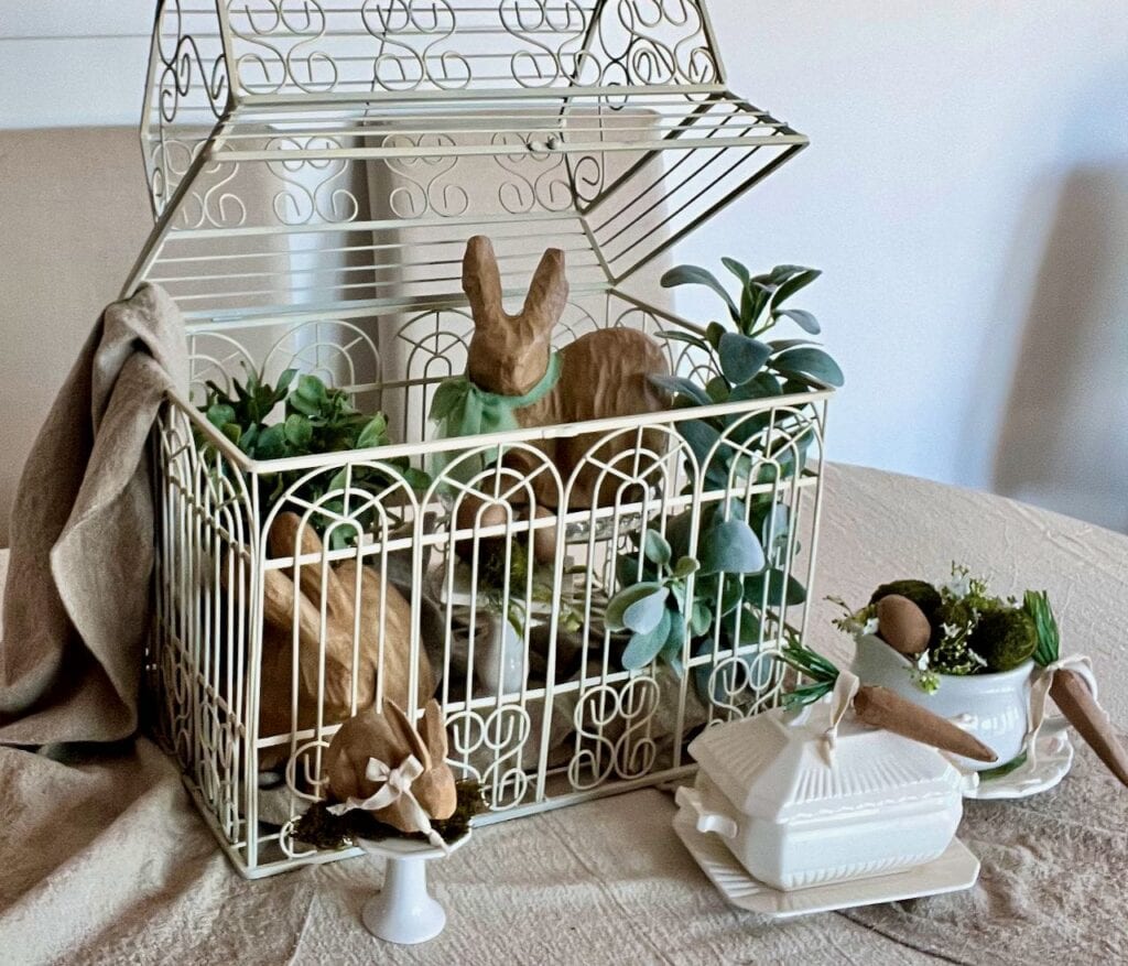 Image of a large decorative birdcage on linen table runners. The lid is propped open with plants, large bunnies, eggs inside and a cupcake plate, tureen and soup mug all coordinating to fill the rest of the buffet table