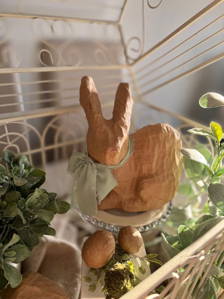 view lookiong down into the decorated birdcage focusing on a large paper mache bunny on a cake plate and soft green plants aruond